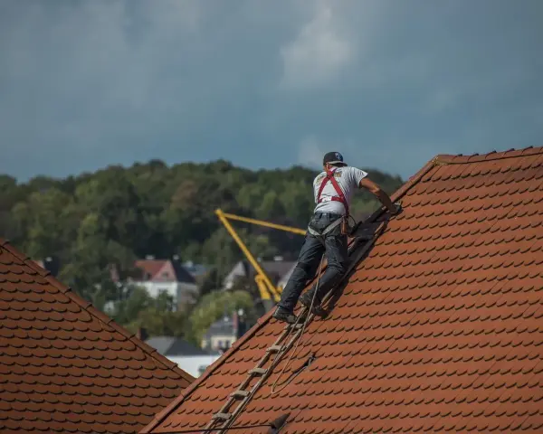 Robotnik w kasku i szelkach bezpieczeństwa na dachu, pracujący przy dachówce.