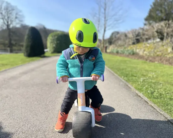 A child in a bright green helmet and teal jacket rides a woom wow balance bike on a sunny path.
