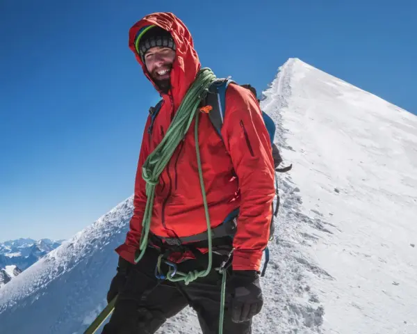 Uśmiechnięty alpinista na szczycie Mont Blanc, w czerwonej kurtce i z liną, gotowy na dalszą wspinaczkę.