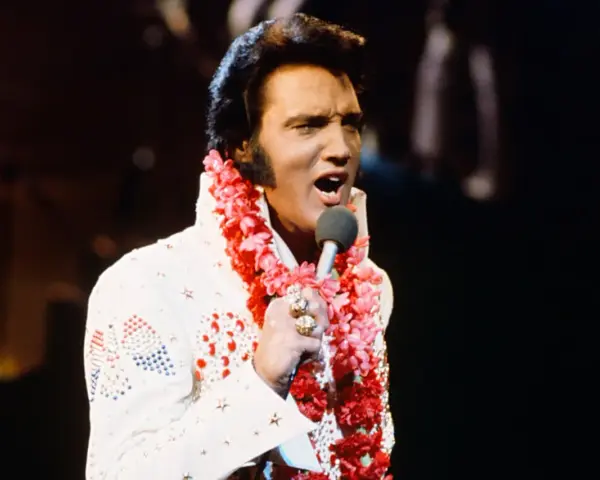Elvis Presley singt in Hawaii, trägt einen weißen Anzug mit Blumenkette und Mikrofon.