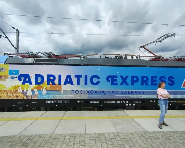 Kobieta stoi obok lokomotywy Adriatic Express z widokiem na morze i żaglówki.