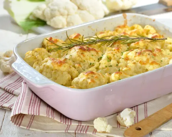 Pyszna keto zapiekanka z kalafiora w różowej naczyniu, udekorowana rozmarynem. Obok świeże różyczki kalafiora.