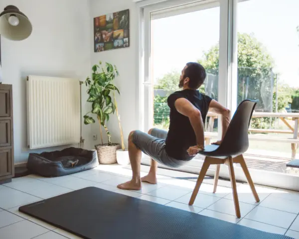 Un homme fait des dips sur une chaise, une idée de quel sport faire à la maison. Un tapis de yoga est au sol.