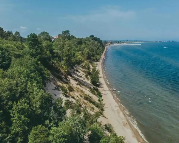 Mechelinki klif porośnięty drzewami, z piaszczystą plażą i turkusowym morzem. W oddali widać zabudowania i falochron.