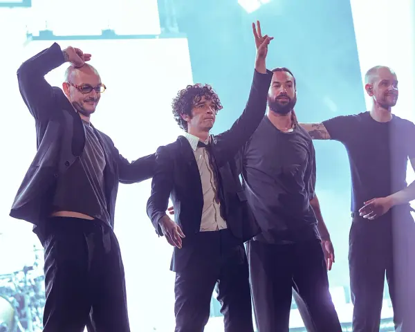 Matty Healy i zespół The 1975 na scenie. Muzycy pozdrawiają publiczność, jeden z nich unosi rękę w geście pokoju.