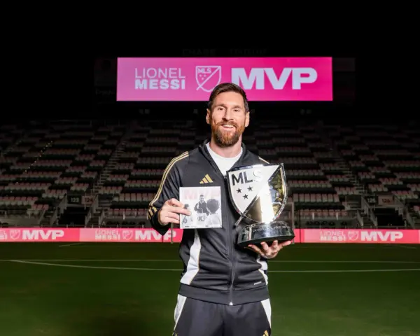 Lionel Messi sonríe en un estadio, sosteniendo un trofeo MLS MVP y un libro. El letrero del estadio dice "LIONEL MESSI MVP".