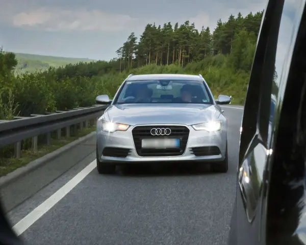 Silberner Audi auf der Autobahn, blendet mit Lichthupe. Vorbei an grünen Wäldern und einer Leitplanke.