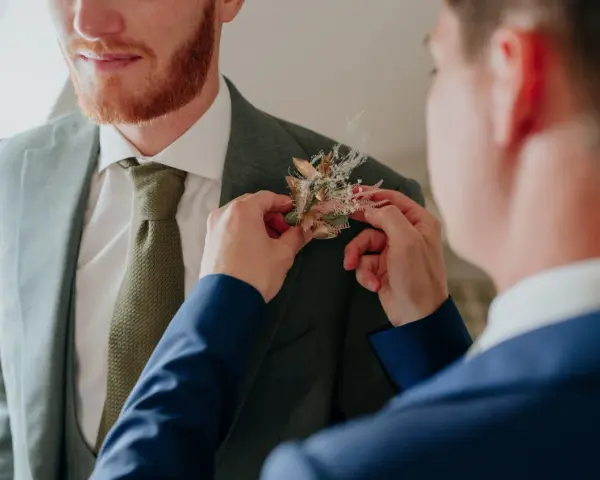 Un homme ajuste une boutonnière sur le costume d'un autre. Divers types de boutonnières existent, celle-ci est composée de fleurs séchées et de feuillages dorés.