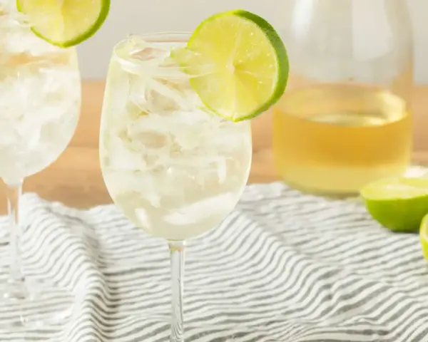 Orzeźwiający szprycer co to? Dwa kieliszki białego wina z lodem i plasterkiem limonki, obok butelka wina i połówki limonki.