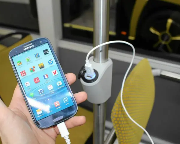 Dłoń trzyma smartfon podłączony do ładowarki USB zamontowanej na słupku w autokarze.