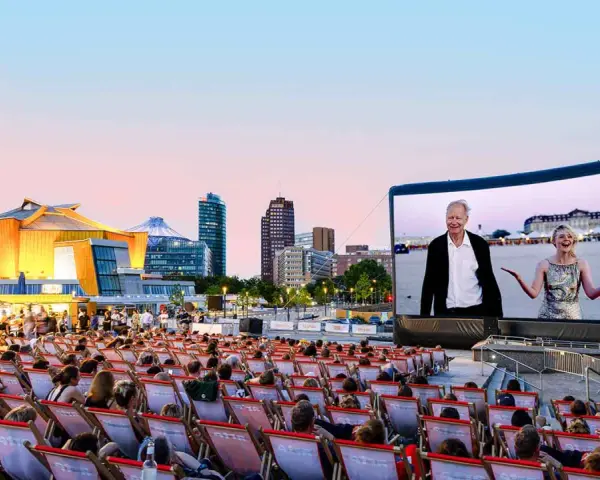 Ein lauer Sommerabend im Freiluftkino Charlottenburg. Menschen sitzen in Liegestühlen und schauen auf eine Leinwand, auf der ein Film mit einem älteren Mann und einer jungen Frau läuft. Im Hintergrund die Berliner Skyline.
