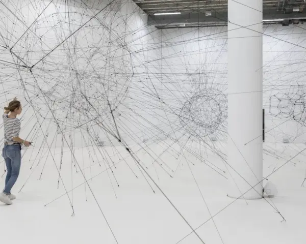 Une femme admire une installation de Tomas Saraceno, un réseau complexe de fils noirs tendus dans un espace blanc.
