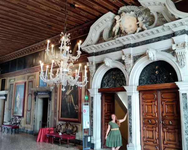 Una donna in un abito verde posa davanti a un portale decorato in una casa museo a Venezia, con un lampadario di cristallo e dipinti alle pareti.