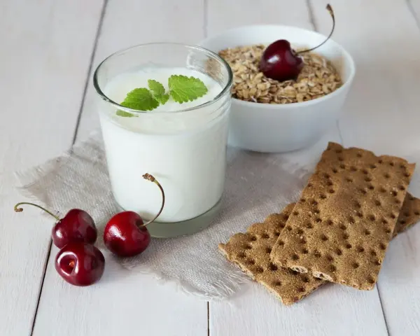 Un verre de yaourt lait végétal, garni de menthe, avec des cerises et des crackers sur une table en bois.