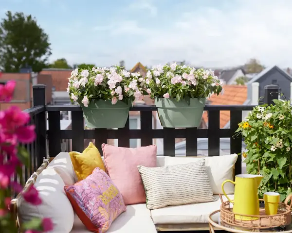 Ein gemütlicher Balkon mit bunten Kissen, gelber Kanne und blühenden sommerblumen.