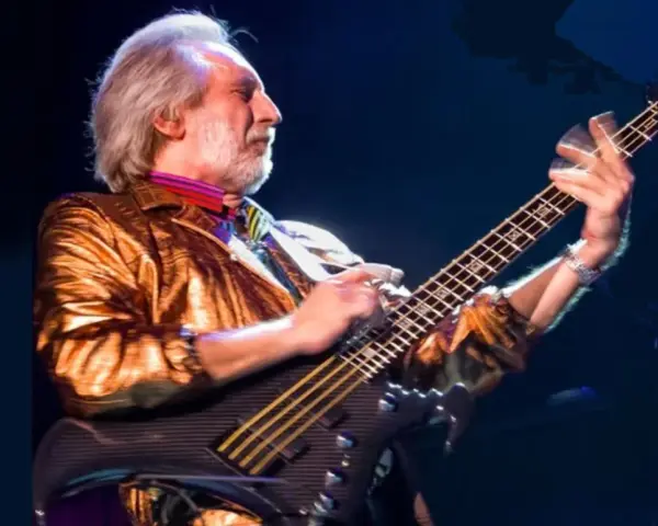 Ein Mann mit grauem Haar und Bart spielt Bassgitarre. Er trägt eine glänzende goldene Jacke und bunte Krawatte. Er ist der beste Bassist der Welt.