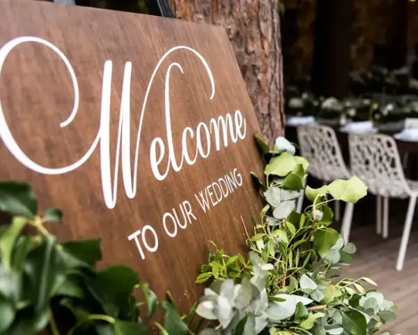 Drewniana tablica "Welcome to our wedding" ozdobiona zielenią. W tle widać przygotowane stoły i krzesła na uroczystość.