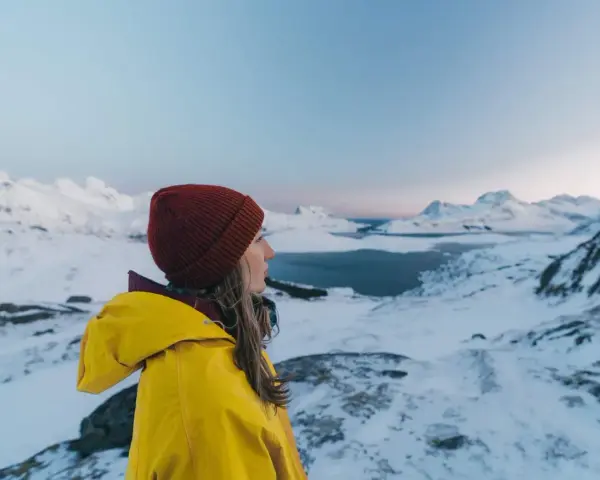 Kvinna i gul jacka och röd mössa njuter av utsikten över snöklädda berg och hav. Perfekt för sportlov i Norge.