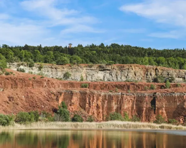En sjö speglar Sveriges Grand Canyon, en imponerande klippformation med lager av röd och grå sten, krönt av en grönskande skog.