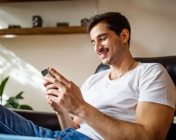Uśmiechnięty mężczyzna w białej koszulce siedzi w fotelu i patrzy na telefon.
