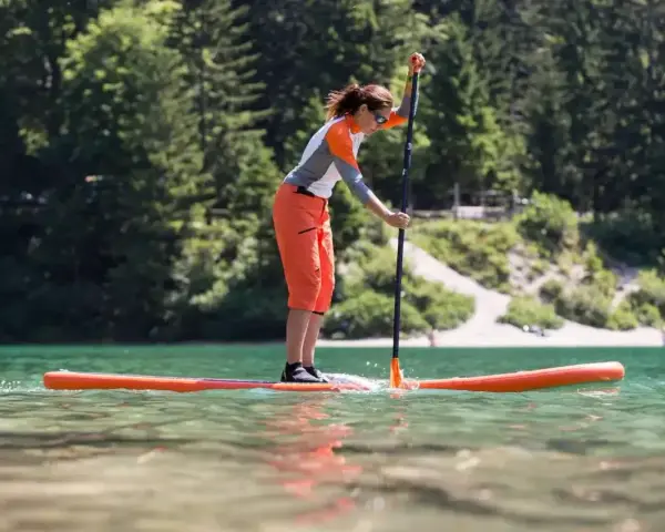 Kobieta w pomarańczowych spodenkach i sportowej bluzce uprawia pływanie na supie po spokojnym jeziorze. W tle zielone drzewa i jasna plaża.
