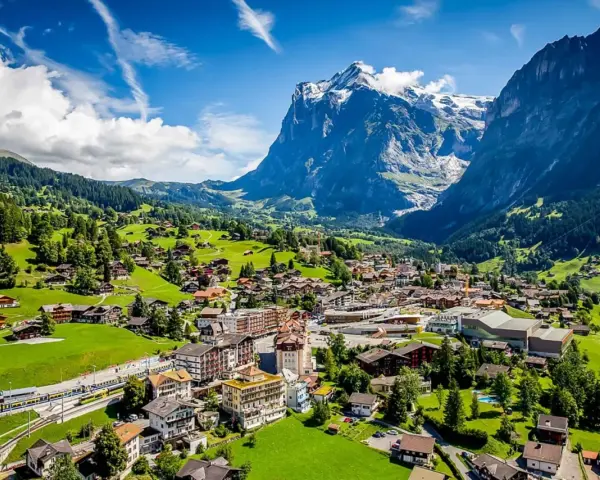 A scenic view of Grindelwald, Switzerland, nestled in a valley with lush green hills and a majestic snow-capped mountain. A train travels through the village.