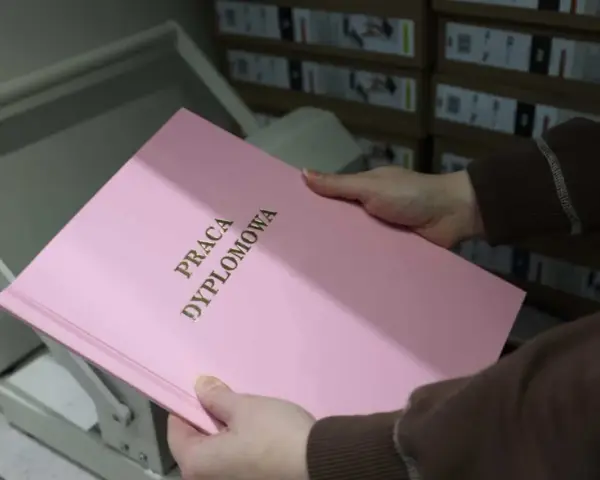 Różowa okładka z napisem "PRACA DYPLOMOWA" w złotych literach, trzymana w dłoniach nad bindownicą.