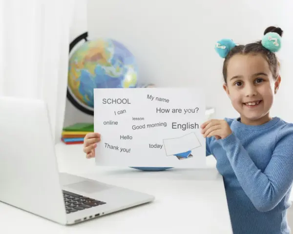 Uśmiechnięta 6latka z tabliczką "English" i zwrotami do nauki. Obok laptop i globus.