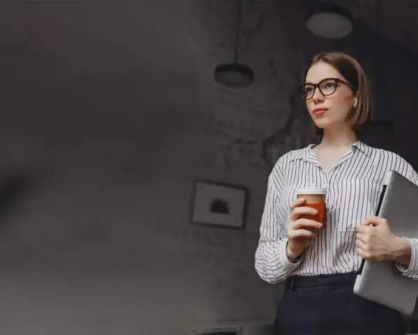 Kobieta w okularach trzymająca kawę i laptopa.