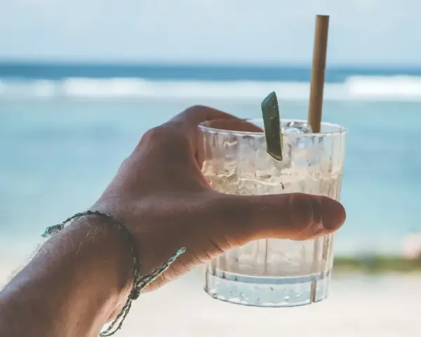 Orzeźwiający drink na plaży, z lodem i plasterkiem limonki, w dłoni z bransoletką. W tle błękitne morze i fale.