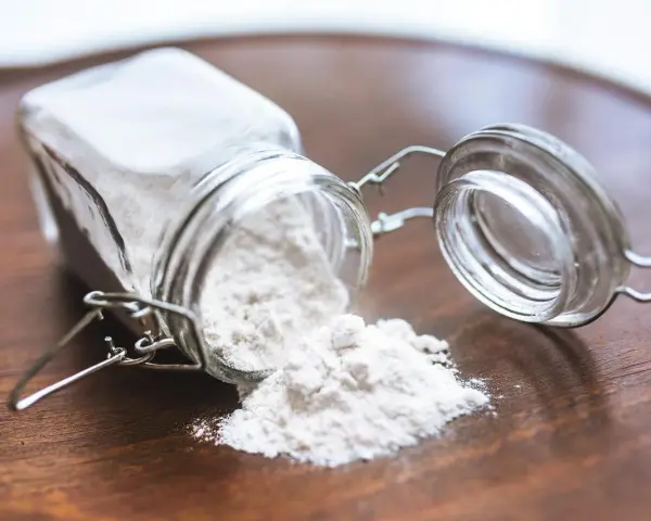 Un barattolo di vetro rovesciato su un tavolo di legno, con polvere bianca che fuoriesce, forse ingredienti per colla vinilica fatta in casa.