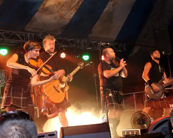 Un groupe celtique français sur scène, avec violon, guitare et basse. Les musiciens portent des kilts, créant une ambiance festive.
