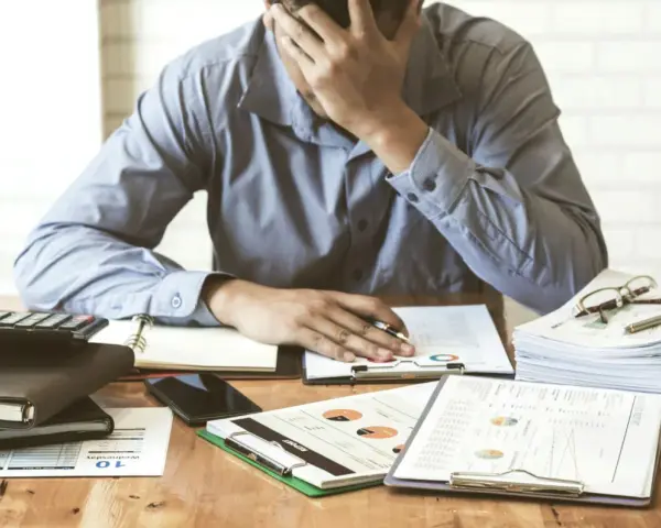 Zestaw dokumentów, kalkulator i telefon leżą na biurku. Mężczyzna z ręką na czole, przytłoczony problemami finansowymi, przegląda pismo windykacyjne.