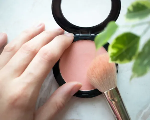 Une main applique un pinceau sur un blush poudre, révélant une douce teinte couleur blush.