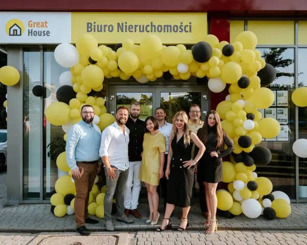 Grupa ludzi pozuje przed biurem nieruchomości ozdobionym żółtymi balonami.