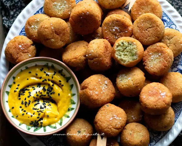 Falafel ricetta originale: croccanti palline dorate, una con un morso rivela il verde interno, accompagnate da una salsa gialla con semi di sesamo nero.