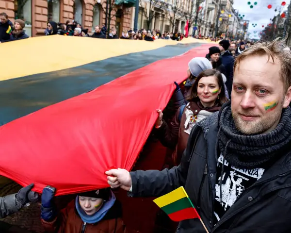 Ludzie niosą ogromną flagę Litwy podczas święta narodowego. Na twarzach uczestników widać patriotyczne barwy.