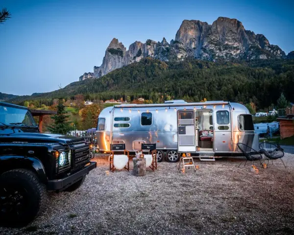 Ein Airstream-Wohnwagen und ein schwarzer Geländewagen stehen auf einem **Campingplatz Dolomiten** mit majestätischen Bergen im Hintergrund. Gemütliche Stühle und Lichter schaffen eine einladende Atmosphäre.