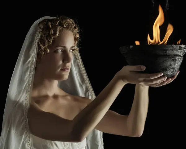 A woman with curly hair and a veil, embodying Hestia goddess, holds a bowl with a sacred flame.