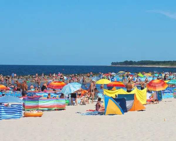 Tłum ludzi na plaży w Darłówku Wschodnim, kolorowe parawany i namioty chronią przed słońcem.