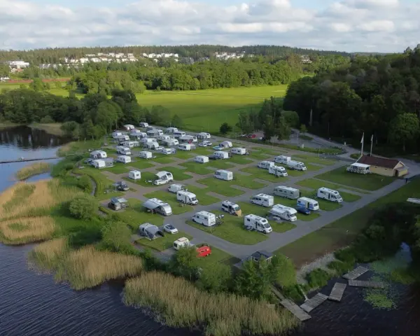 Full campingplats med husbilar och husvagnar vid en sjö, med grönskande träd och bebyggelse i bakgrunden. En idyllisk plats för camping i Alingsås.