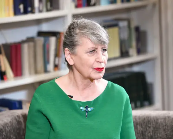 Joanna Wnuk-Nazarowa w zielonej bluzce z broszką w kształcie ważki, na tle regałów z książkami.