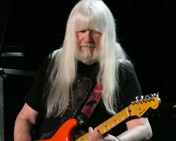 Andy Scott mit seinen charakteristischen langen, weißen Haaren spielt auf einer orangen E-Gitarre.
