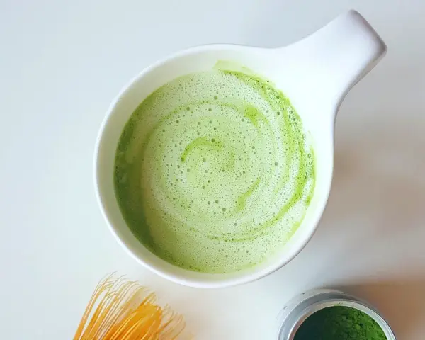 Zielony, spieniony napój matcha w białej miseczce, obok bambusowa miotełka i puszka z proszkiem. Idealne co na pobudzenie zamiast kawy.