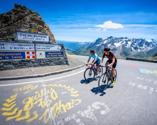 Ciclisti in salita verso il Col du Galibier, con montagne innevate sullo sfondo e segnaletica stradale.