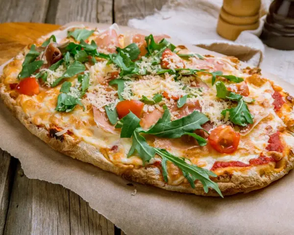Pinsa co to? Aromatyczna, podłużna pizza z rukolą, pomidorkami i szynką parmeńską, posypana parmezanem.