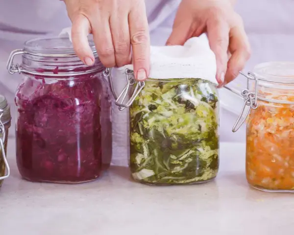 Hände bedecken ein Glas mit fermentiertem Gemüse, Teil einer Auswahl an probiotischen Lebensmitteln in Gläsern.