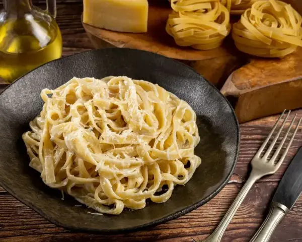 Pyszne Fettuccine Alfredo przepis na talerzu, obok surowy makaron, ser i oliwa. Idealne danie dla miłośników włoskiej kuchni.