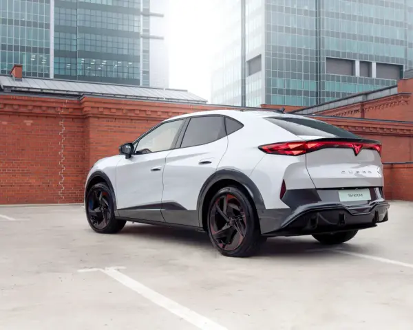 Biały elektryczny SUV Cupra Tavascan z czerwonymi akcentami na felgach, zaparkowany na tle ceglanego muru i nowoczesnych budynków.