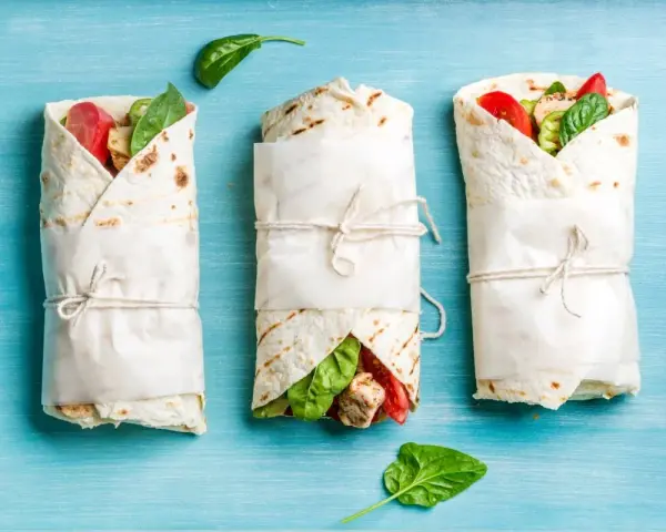 Trzy keto tortille z siemienia lnianego, zawinięte w papier i przewiązane sznurkiem, z widocznym nadzieniem z kurczaka, pomidorów i szpinaku.